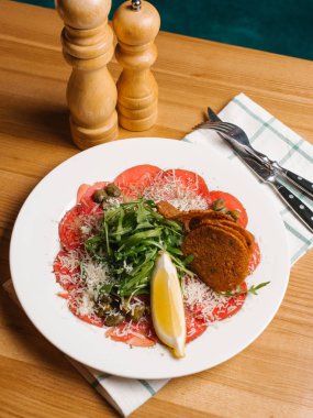 Biberli ekmek tost ile dilimlenmiş, roka, Kapari ve limon dilimi ile rendelenmiş parmesan, bıçak, çatal, tuz dekore edilmiş beyaz tabakta servis ve öğütücüler ve Damalı peçete biber