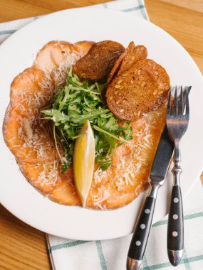 Dilimlenmiş ekmek tost ile somon füme, roka ve limon dilim rendelenmiş parmesan ile dekore edilmiş bıçak, çatal ve Damalı peçete beyaz tabakta