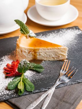 Cheesecake karamel şurubu ile dilimlenmiş çilek, nane yaprakları ve pudra şekeri, çatal, çaydanlık ve bardak çay ile taş tabakta servis