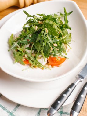 Tabak, çatal, bıçak, karabiber öğütücüsü ve peçete ahşap masa üzerinde beyaz kase dilimlenmiş domates ve çam fıstığı roka, kızarmış yumurta closeup hizmet