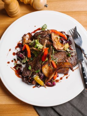 Kavrulmuş rack kuzu kırmızı biber, karamelize tatlı soğan, marul ve bezelye lahanası ile görünümünü peçete, çatal, bıçak, biber ve tuz değirmenleri ile beyaz tabakta servis