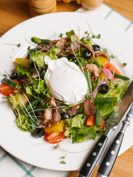 Salad with smoked eel, sweet peppers, cherry tomatoes, olives, lettuce, sweet onion, green beans and peas sprouts served on white plate with yoghurt, knife, fork, salt and pepper grinders