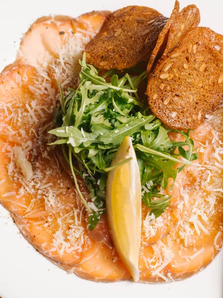 Sliced smoked salmon with bread toasts, arugula and lemon slice served on white plate decorated with grated parmesan