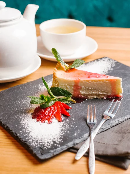 Cheesecake with fruit syrup served on stone plate with sliced strawberry, mint leaves and forks, teapot and cup of tea