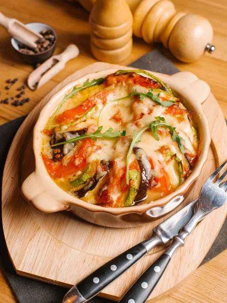 Baked eggplants, zucchini and tomatoes with cheese and fresh arugula served in ceramic bowl on wooden chopping board with fork and knife