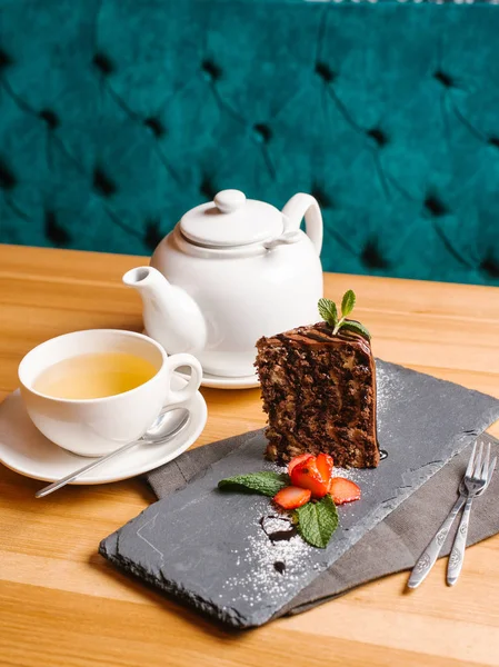Chocolate zebra cake served on stone plate with sliced strawberry, mint leaves, powdered sugar, cup of tea, teapot, forks and spoon on wooden table