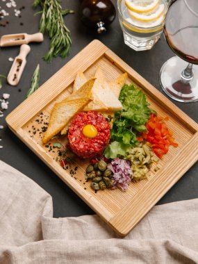 Biftek tartare ile Bıldırcın yumurta, ekmek tost, doğranmış turşu, tatlı soğan, domates, marul ve Kapari servis ahşap tabak üzerinde bıçak, çatal, şarap ve limonata ile