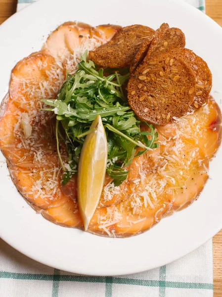 Sliced smoked salmon with bread toasts, arugula and lemon slice served on white plate decorated with grated parmesan