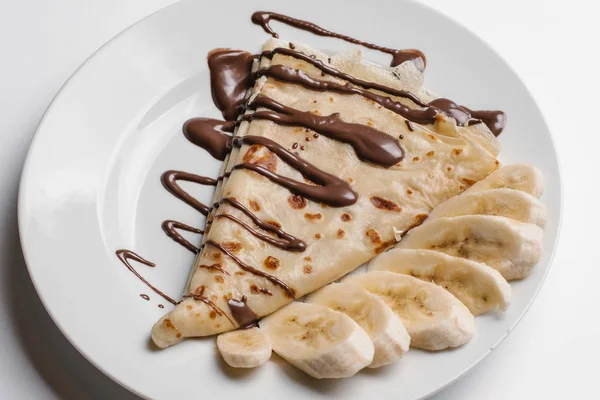 Closeup of crepe with chocolate syrup and sliced banana served on white plate isolated on white background