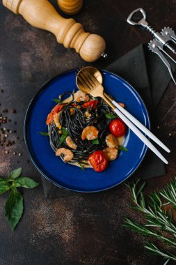 Masada karides, domates ve yeşillik çatal ve kaşık, karabiber öğütücüsü, şişe açacağı ve otlar ile mavi plaka üzerinde siyah makarna Görünümü Kapat