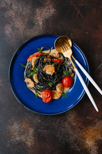 Black pasta with shrimps, tomatoes and greenery on blue plate with fork and spoon