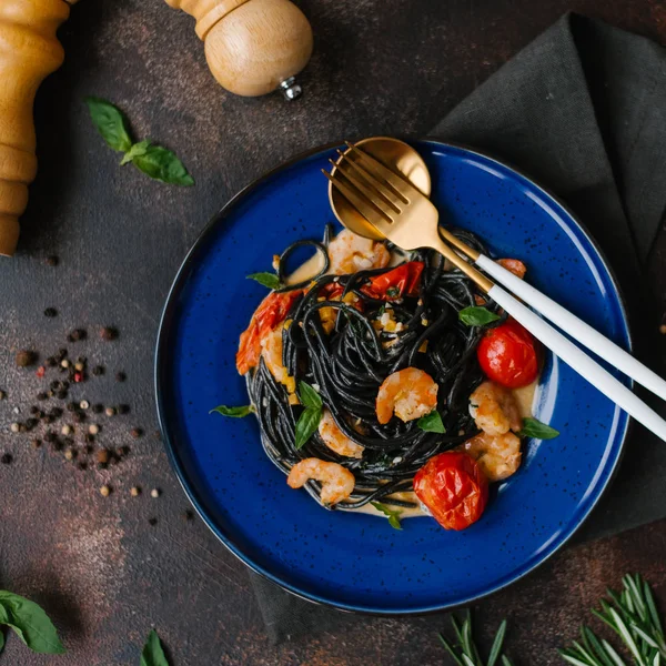 Closeup of black pasta with shrimps, tomatoes and greenery on blue plate with fork and spoon, pepper grinder and herbs on table