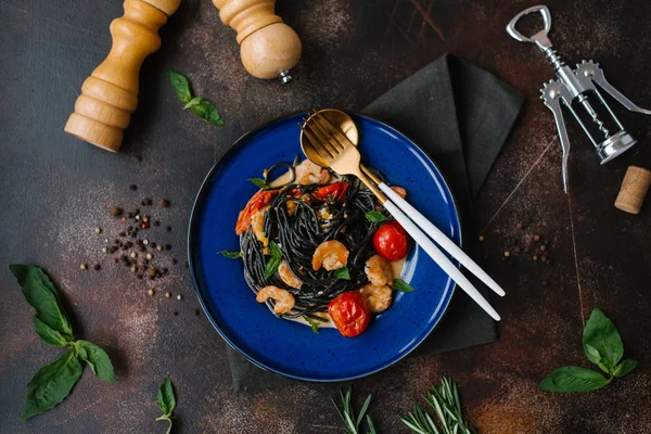 Close view of black pasta with shrimps, tomatoes and greenery on blue plate with fork and spoon, pepper grinder, bottle opener and herbs on table