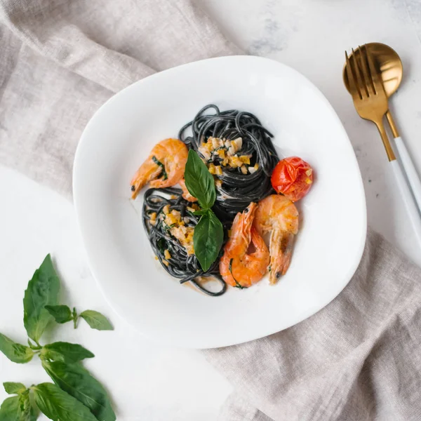 Black pasta with shrimps, tomatoes and greenery on white plate with napkin, fork and spoon