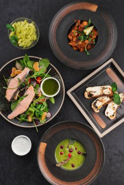 set of dishes with baked fish, green salad, broccoli cream soup and yogurt and pesto sauces on black background 