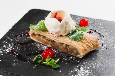 Apple strudel decorated with scoop of ice cream, cherries and sugar powder served on black plate