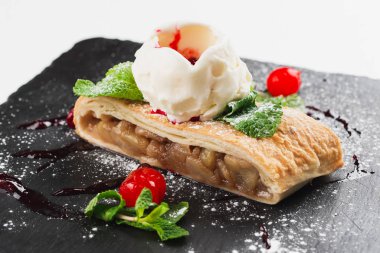 Apple strudel decorated with scoop of ice cream, cherries and sugar powder served on black plate
