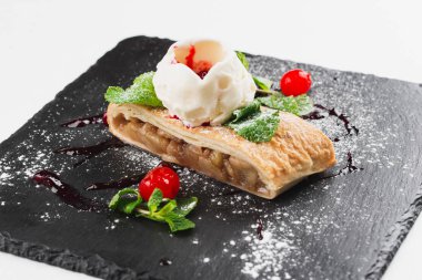 Apple strudel decorated with scoop of ice cream, cherries and sugar powder served on black plate
