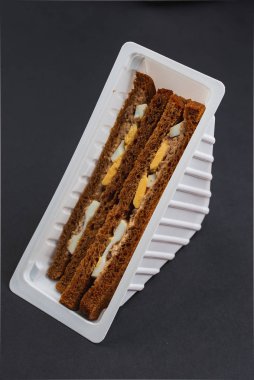black triangular bread sandwiches with chicken, cheese and eggs in plastic box on the black table 