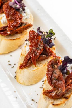 Krem peynirli beyaz ekmek dilimleri ve zeytinyağlı kurutulmuş domatesler ve beyaz tabakta otlar.