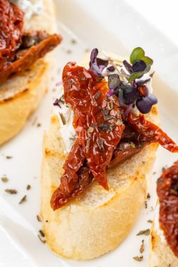 Krem peynirli beyaz ekmek dilimleri ve zeytinyağlı kurutulmuş domatesler ve beyaz tabakta otlar.