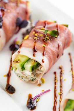 Parma jambonlu kanepe, yeşil salatalıklı ve beyaz tabakta kahverengi soslu.  