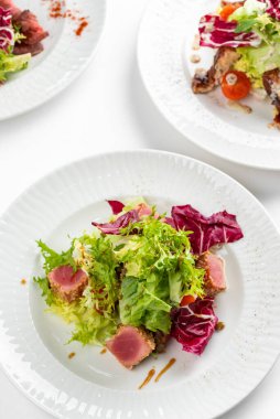Beyaz masa arkasında ton balığı dilimleri, somon, yeşillik ve domatesli renkli salata tabakları.