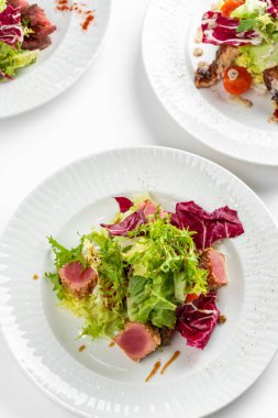 Beyaz masa arkasında ton balığı dilimleri, somon, yeşillik ve domatesli renkli salata tabakları.