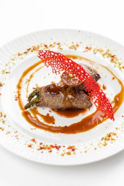 Kahverengi sosun altında yeşil kuşkonmaz ve beyaz tabakta kırmızı krakerle süslenmiş fırınlanmış et ruloları. 