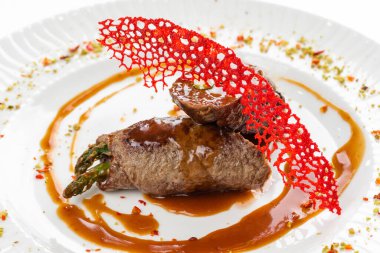 Kahverengi sosun altında yeşil kuşkonmaz ve beyaz tabakta kırmızı krakerle süslenmiş fırınlanmış et ruloları. 