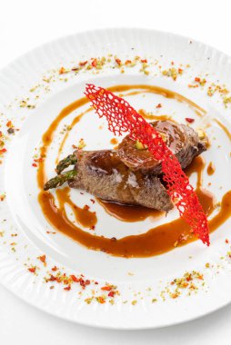 Kahverengi sosun altında yeşil kuşkonmaz ve beyaz tabakta kırmızı krakerle süslenmiş fırınlanmış et ruloları. 