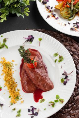 Kırmızı sosun altında kızarmış ördek fileto ve beyaz tabakta domatesli sebze tabakları.