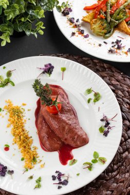 Kırmızı sosun altında kızarmış ördek fileto ve beyaz tabakta domatesli sebze tabakları.