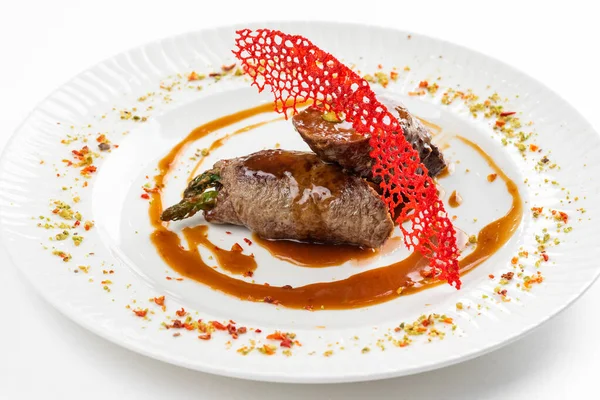 Kahverengi sosun altında yeşil kuşkonmaz ve beyaz tabakta kırmızı krakerle süslenmiş fırınlanmış et ruloları. 