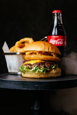 Siyah masa arka planında metal sepetin içinde Coca Cola cam şişe ve kızarmış soğan halkaları ile etli burger.