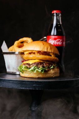 Siyah masa arka planında metal sepetin içinde Coca Cola cam şişe ve kızarmış soğan halkaları ile etli burger.