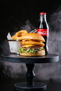 Siyah masa arka planında metal sepetin içinde Coca Cola cam şişe ve kızarmış soğan halkaları ile etli burger.