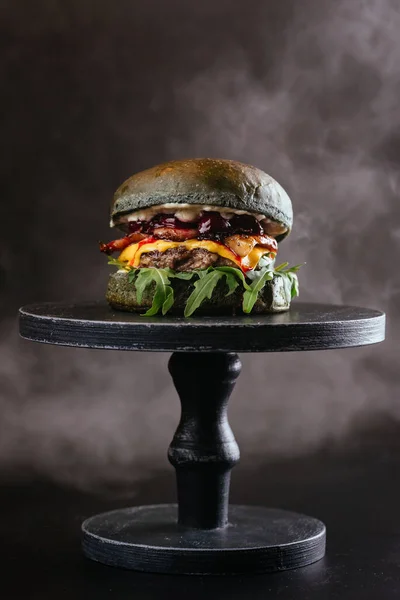 black buns meat burger with cheese, red cherries sauce and green arugula leaves on wooden table background