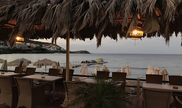 Wieczorny śródziemnomorski krajobraz z morzem, zatoką, plażą, parasolami, palmami, stołami, krzesłami i oświetlonymi lampami — Zdjęcie stockowe