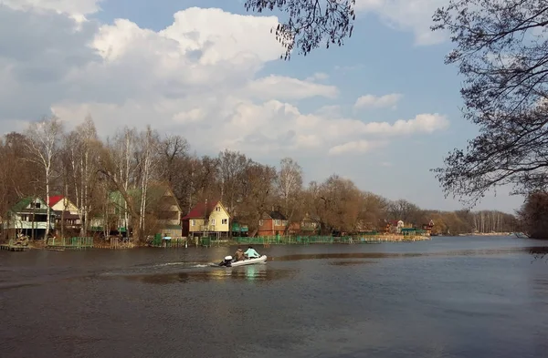 Paesaggio primaverile con un grande fiume, un villaggio sulla riva, case rurali colorate e motoscafo galleggiante sul fiume con persone — Foto Stock