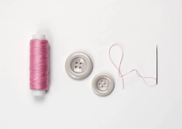 Spool of pink thread, needle, two buttons on a white background.