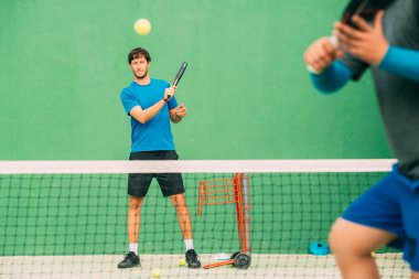 Genç adam yeşil bir kortta raket tenis oynuyor, yeşil bir kortta blueYoung tenis oynuyor, mavi bir tişört giyiyor.