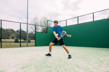 Yeşil kortta raket oynayan genç bir adam, mavi tişört giyiyor. Uzay konseptini kopyala.