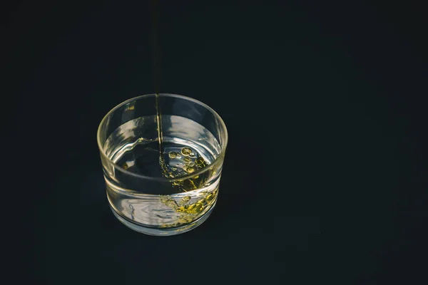 Olio d'oliva che cade sull'acqua in un bicchiere basso con gocce d'olio sul fondo . — Foto Stock