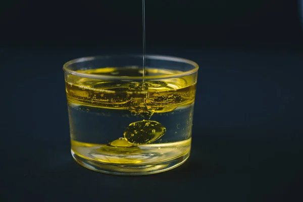 Olio d'oliva che cade sull'acqua in un bicchiere basso con gocce d'olio sul fondo — Foto Stock