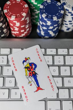 Arkaplanda renkli çip kolonları olan bir bilgisayar klavyesinde Joker poker kartı. Yukarıdan görüntü. Şans kavramı, bahis ve online oyun