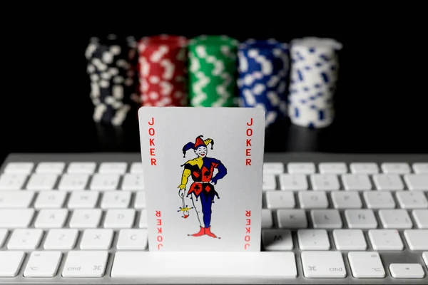 JOKER poker card on a computer keyboard with poker chips placed in columns unfocused on the background.Isolated on black background.Concept of chance, betting and online game