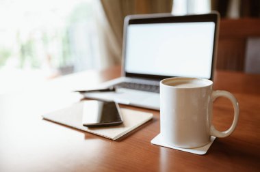 Merkez ofis masası. Laptoplu beyaz kahve fincanı ve ahşap masada akıllı telefon. Gün ortası ışığı pencereden içeri giriyor. Seçici odaklan. İş ve ev kavramı