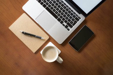 Ofisteki ahşap masa, dizüstü bilgisayarı, beyaz kahve fincanı ve akıllı telefonu olan. Yukarıdan görüntü eğimli. İş, iletişim ve teknoloji kavramı