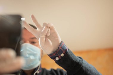 Evdeki karantina sırasında elinde cep telefonuyla zafer işareti yapan bir adam. Grip maskesi ve hijyenik eldivenler takmak. Seçici odaklanma. Covid19 konseptine göre ev hapsi.
